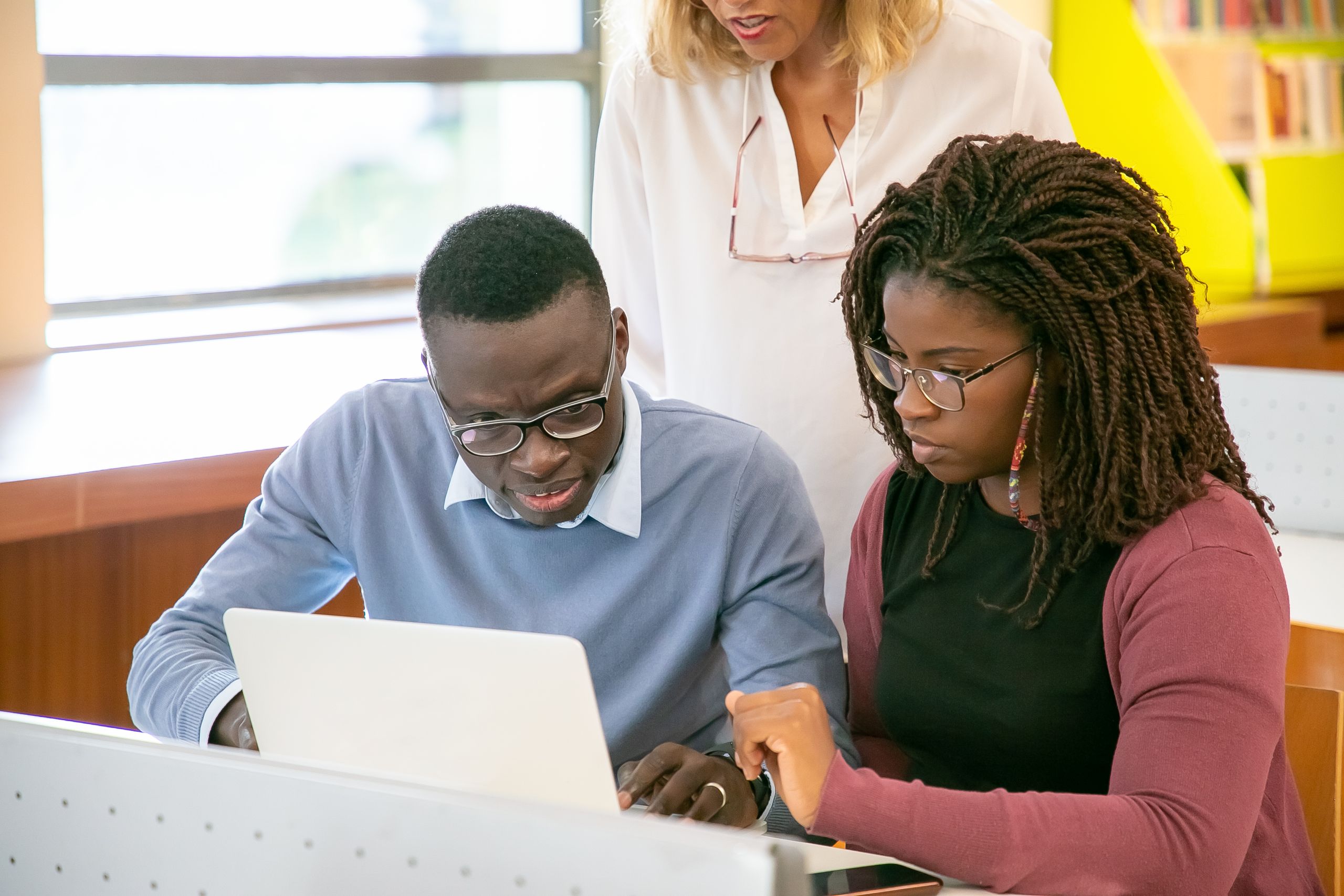 Crop unrecognizable female tutor in white shirt helping young African American students in eyeglasses doing research using laptop in light classroom